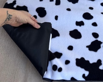 May include: A black and white cow print blanket with a black backing. The blanket features large, irregular black spots on a white background. The corner of the blanket is folded over, revealing the black backing material.