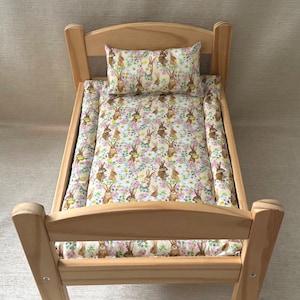 Op de afbeelding: Een houten poppenbed met een wit en bruin beddengoedset met konijnen en bloemen. Het beddengoed omvat een kussen en een hoeslaken.