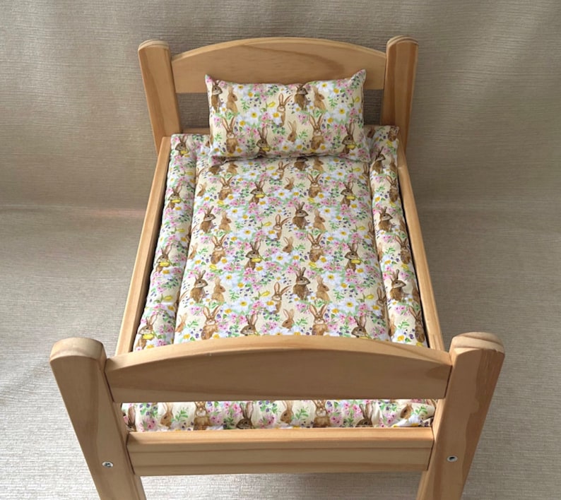 Op de afbeelding: Een houten poppenbed met een wit en bruin beddengoedset met konijnen en bloemen. Het beddengoed omvat een kussen en een hoeslaken.