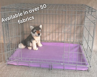 May include: A metal pet crate with an open door, a purple bed, and a stuffed wolf toy inside. The text "Available in over 50 fabrics" is in the upper left corner. The crate is designed for pets.
