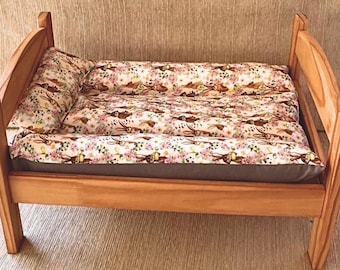 May include: A small wooden bed frame with a light brown finish, featuring a mattress and a pillow covered in a cream-colored fabric with a repeating pattern of brown rabbits, pink flowers, and green leaves, set on a dark gray base.