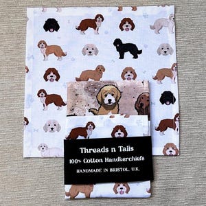 Może przedstawiać: Dwa białe chusteczki z bawełny z powtarzającym się wzorem brązowych i białych psów. Górna chusteczka ma białą obwódkę z niebiesko-białym wzorem. Dolna chusteczka ma białą obwódkę z brązowo-białym wzorem. Chusteczki mają napis "Threads n Tails 100% Cotton Handkerchiefs Handmade in Bristol, U.K."