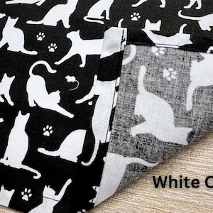 May include: Black fabric with a repeating pattern of white cat silhouettes, paw prints, and small mouse illustrations. The fabric is folded over, revealing a lighter, similar pattern. The text "White Cats" is visible.