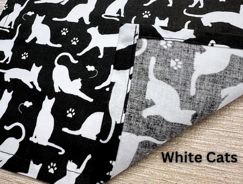 May include: Black fabric with a repeating pattern of white cat silhouettes, paw prints, and small mouse illustrations. The fabric is folded over, revealing a lighter, similar pattern. The text "White Cats" is visible.