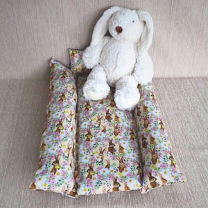 Könnte beinhalten: Ein weißes Plüschkaninchen sitzt auf einem langen, rechteckigen Kissen mit einem Blumenmuster mit braunen Kaninchen. Das Kissen besteht aus zwei separaten Teilen.