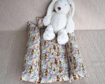 May include: A white plush bunny toy sits on a long, rectangular pillow with a floral pattern featuring brown bunnies. The pillow is made of two separate pieces.