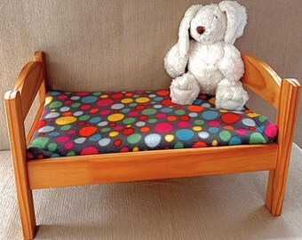 May include: A small wooden pet bed with a colorful polka dot patterned cushion. A white stuffed bunny sits on the bed. The bed frame is made of light-colored wood.