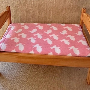May include: A small wooden bed frame with a natural light brown finish, featuring arched headboard and footboard panels. A soft rectangular blanket with a pink background and a repeating white rabbit pattern is placed flat across the bed frame.