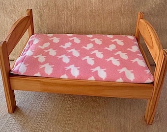 May include: A small wooden bed frame with a natural light brown finish, featuring arched headboard and footboard panels. A soft rectangular blanket with a pink background and a repeating white rabbit pattern is placed flat across the bed frame.