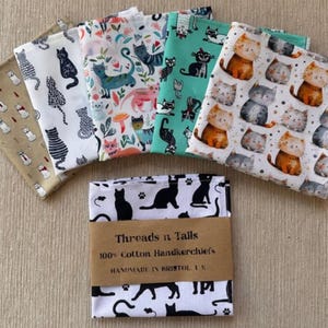 May include: A collection of folded cotton handkerchiefs with various cat-themed patterns. Designs include black cats, cartoon cats, and cats with floral elements. The bottom handkerchiefs are wrapped with a brown paper band that reads "Threads n Tails".