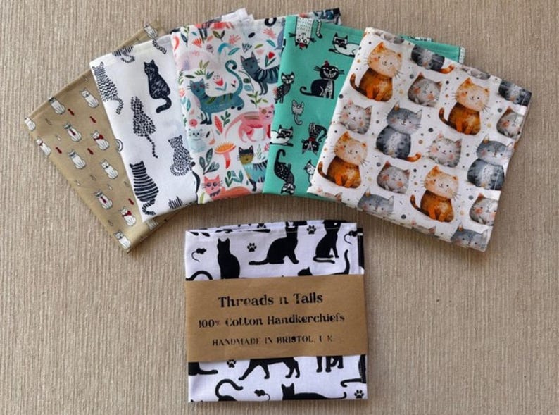 May include: A collection of folded cotton handkerchiefs with various cat-themed patterns. Designs include black cats, cartoon cats, and cats with floral elements. The bottom handkerchiefs are wrapped with a brown paper band that reads "Threads n Tails".