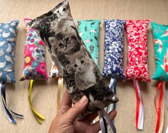 May include: A collection of rectangular fabric cat toys featuring various patterns including kittens, floral designs, and colorful motifs. Each toy is finished with a set of dangling satin ribbons at the bottom, showcasing diverse prints like blue, red, and teal.