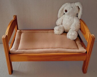 May include: A small wooden bed with a light brown cushion and a white stuffed bunny. The bed frame is made of light-colored wood. The bunny has long ears and a brown nose.