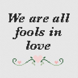 May include: Cross-stitch design on white fabric with the words "We are all fools in love" in black. A decorative border of green vines, pink flowers, and a pink heart is at the bottom. The design has a handmade, vintage aesthetic.