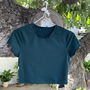 K&ouml;nnte beinhalten: Ein dunkelgr&uuml;nes, &auml;rmelloses, bauchfreies T-Shirt mit Rundhalsausschnitt. Das Shirt h&auml;ngt an einem wei&szlig;en Kleiderb&uuml;gel.