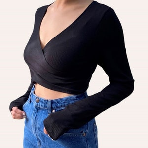 K&ouml;nnte beinhalten: Ein schwarzes, lang&auml;rmliges Wickel-Crop-Top mit tiefem V-Ausschnitt. Das Oberteil ist aus geripptem Stoff und hat eine figurbetonte Silhouette.