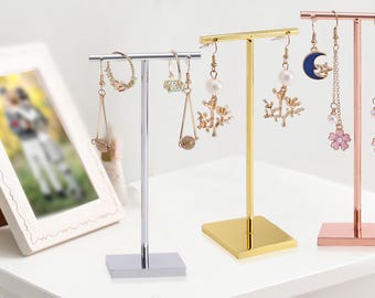 May include: Three earring display stands in silver, gold, and rose gold. Each stand has a T-shaped bar for hanging earrings. The stands hold various earrings, including hoops, drop earrings, and dangle earrings with pearls, floral, and celestial designs.