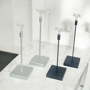 May include: Three display stands for merchandise. Two are silver and one is black. Each stand has a clear plastic clip at the top and a square base.