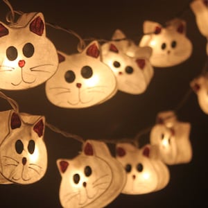 Pode incluir: Uma corda de lanternas de papel brancas em forma de gato com pequenas lâmpadas dentro. As lanternas são amarradas com um fio transparente e estão acesas, criando um brilho quente.