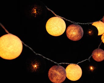 Puede incluir: Guirnalda de luces con múltiples bolas esféricas de algodón tejido en tonos naranja, marrón y negro. Las luces están ensartadas en un fino cable blanco sobre un fondo oscuro, creando un ambiente cálido y acogedor.