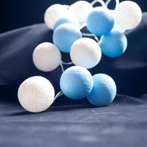 May include: A string of white and blue cotton ball lights. The lights are strung together with a clear wire and are hanging on a dark blue fabric background.