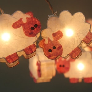 Könnte beinhalten: Lichterkette aus weißen Papierschafen mit rosafarbenen Gesichtern und roten Hufen. Die Lichter sind an und leuchten.