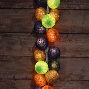 May include: A string of cotton ball lights with a mix of green, orange, and blue lights. The lights are strung together with a white wire and are hanging against a wooden background.