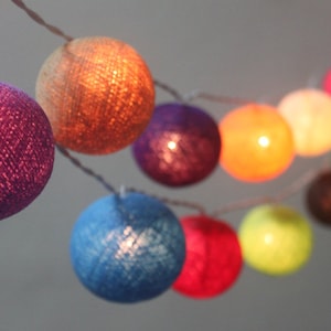 May include: A string of colourful cotton ball lights. The lights are strung together with a clear wire and are lit with warm white LED bulbs. The cotton balls are in various colours, including purple, orange, blue, green, red, and yellow.