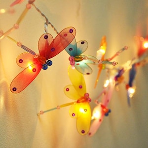 Puede incluir: Una cadena de luces de hadas con bombillas de colores en forma de libélula. Las luces están enhebradas en un cable transparente y están encendidas, proyectando un brillo cálido.