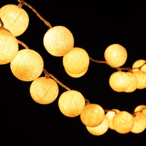 May include: A string of white cotton ball lights with warm white LED bulbs. The lights are strung together with a clear wire and are hanging against a black background.