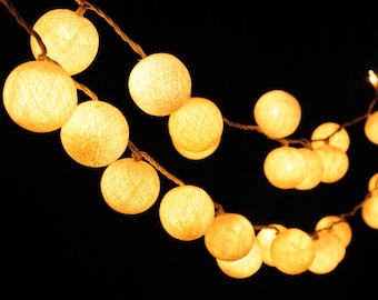 May include: A string of white cotton ball lights with warm white LED bulbs. The lights are strung together with a clear wire and are hanging against a black background.