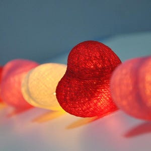 Könnte beinhalten: Eine Lichterkette aus Baumwollkugeln mit einem roten herzförmigen Licht in der Mitte. Die Lichter sind in einer Reihe aneinandergereiht und leuchten, wodurch ein warmes Licht entsteht.
