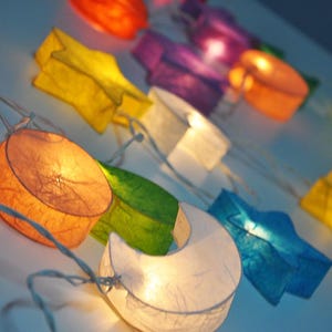Puede incluir: Una cadena de luces de papel de colores con forma de estrellas y lunas. Las luces están enhebradas en un cable blanco y están encendidas, creando un brillo cálido.