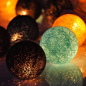 Pode incluir: Uma corda de luzes decorativas com bolas de algodão em várias cores, incluindo preto, laranja, azul-petróleo e marrom. As luzes estão acesas e brilhando.