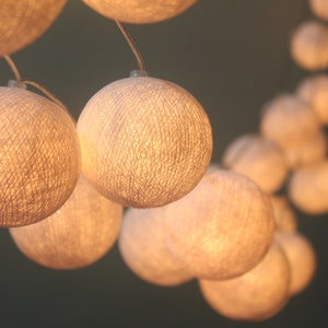 Puede incluir: Primer plano de una cadena de luces esféricas brillantes. Las luces están hechas de un material tejido, emitiendo un brillo cálido y suave. El fondo es verde apagado, creando contraste. Ideal para la decoración del hogar.