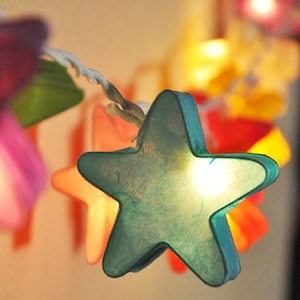 Pode incluir: Cordão de luzes com lanternas de papel em forma de estrela em várias cores, incluindo turquesa, rosa e amarelo. As luzes estão acesas e brilhando.