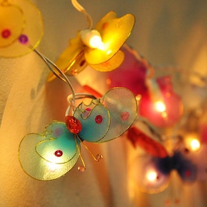 Pode incluir: Uma corda de luzes com lâmpadas em forma de borboleta. As lâmpadas são feitas de tecido translúcido e vêm em várias cores, incluindo amarelo, azul, rosa e verde. Cada lâmpada tem uma pequena luz dentro, criando um brilho quente.
