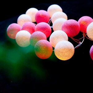 May include: A string of cotton ball lights with a mix of white and pink lights. The lights are strung together with a clear wire.