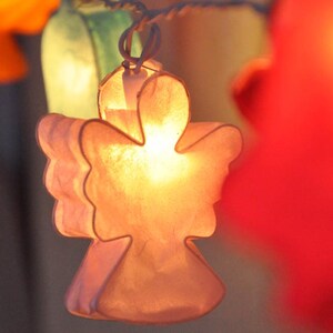Pode incluir: Uma corda de lanternas de papel com uma luz dentro de cada uma. As lanternas têm a forma de anjos e são feitas de um material translúcido. As lanternas estão penduradas em uma corda e são iluminadas por dentro.