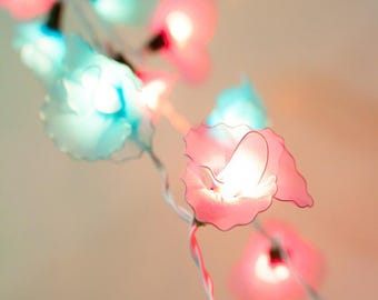May include: A string of flower-shaped fairy lights in shades of pink and blue. The lights are strung on a thin, white wire and emit a soft, warm glow. The background is a blurred gradient of light colors.