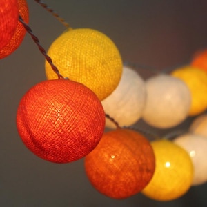 May include: A string of cotton ball lights with a mix of orange, yellow, and white colors. The lights are strung together with a clear wire.
