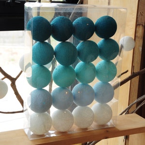 Op de afbeelding: Een set van 20 katoenen ballampjes in een doorzichtige plastic verpakking. De lampjes zijn gerangschikt in drie rijen van zes, waarbij de bovenste rij teal is, de middelste rij lichtblauw en de onderste rij wit.
