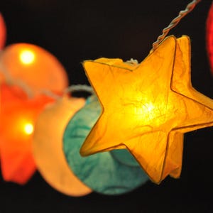 Puede incluir: Cadena de luces con faroles de papel con forma de estrellas y lunas. Los faroles están iluminados con luces blancas cálidas y son de varios colores, incluyendo amarillo, naranja, azul y rojo.