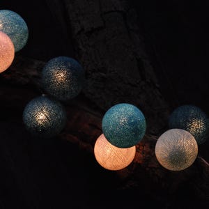 May include: A string of teal and white cotton ball lights hanging from a tree branch. The lights are lit and glowing.