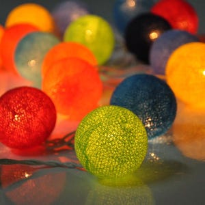 May include: A string of colorful cotton ball lights. The lights are in various colors, including orange, red, blue, green, and yellow. The lights are strung together with a clear wire.