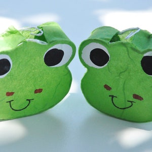 May include: Two green frog-shaped gift boxes with large black eyes, white eye surrounds, and smiling mouths. The boxes have small red cheek accents and are made of a textured material. The boxes are side by side.