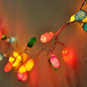 May include: A string of colourful lights with paper lanterns in various shapes and colours. The lights are strung on a wire and are hanging from a wall.