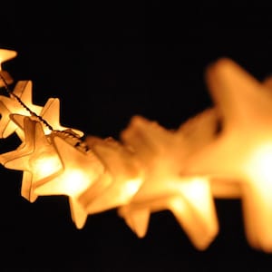 Puede incluir: Una cadena de luces blancas en forma de estrella, brillando intensamente sobre un fondo oscuro. Las luces están conectadas por un cable delgado y crean un ambiente festivo y caprichoso.