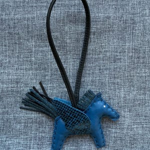 May include: A blue leather Pegasus charm with black wings and a black leather strap. The charm has a detailed design and is set against a gray textured background. The Pegasus has a small white dot for an eye.