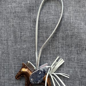 May include: A brown leather horse-shaped charm with a silver strap. The horse has a snakeskin-patterned wing and white fringe. The charm is likely a keychain or bag accessory.